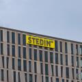 The top corner of the modern Stedin office building, showing its distinctive facade and the company's bright yellow logo against a grey sky. Rotterdam