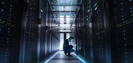 IT Technician Works on Laptop in Big Data Center full of Rack Servers. He Runs Diagnostics and Maintenance.