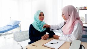 A young beautiful Muslim doctor is using a stethoscope to listen to the patient's heartbeat and write a report at the table in the hospital examinatio