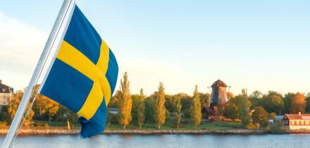 Swedish flag waving over calm waters in the Stockholm archipelago, with Djurgarden island and autumn foliage in the background on a peaceful early Oct