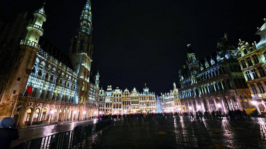 Night view of the illuminated Grand Place in Brussels, Belgium, with historic buildings and crowds gathered in the square.