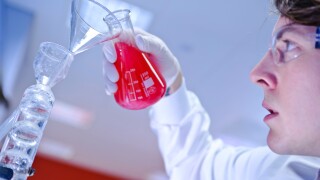 Young make scientist wearing goggles gloves and lab coat pouring bright red liquid from glass beaker into in science laboratory