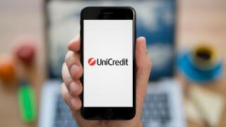 A man looks at his iPhone which displays the UniCredit logo, while sat at his computer desk (Editorial use only).