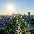 Sweeping aerial view along Avenue de la Grande Armee in Paris, framed by classic Haussmann buildings and green trees, with the modern towers of La Defense silhouetted against a golden evening sky.