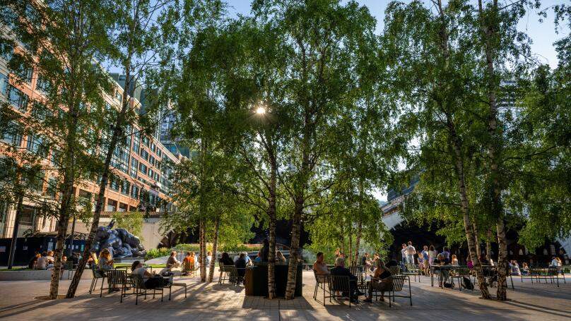 London, UK: Exchange Square at Broadgate, an open space in the City of London. People sit in the square and evening sunlight filters through the trees