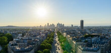 Sweeping aerial view along Avenue de la Grande Armee in Paris, framed by classic Haussmann buildings and green trees, with the modern towers of La Defense silhouetted against a golden evening sky.