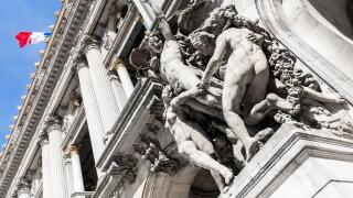 Close up of a group of marble statues on the facade of a french building, with a french flag waving in the background