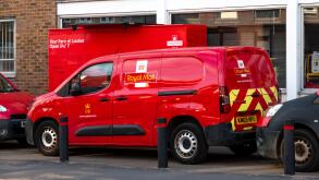 A red Royal Mail delivery van is parked in front of a parcel locker in Bishops Stortford, Hertfordshire, England. The van displays the Royal Mail logo