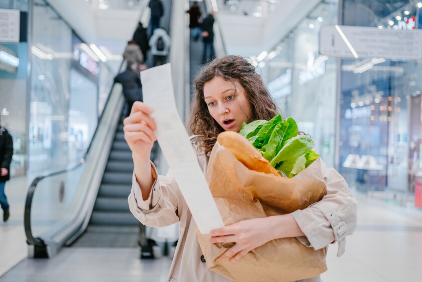 A woman with a paper bag with groceries stands in a shopping center and looks in surprise at a check with high prices.