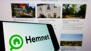 Mobile phone with logo of Swedish property platform company Hemnet AB on screen in front of business website. Focus on center-left of phone display.