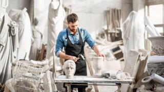 Portrait of a handsome sculptor in blue t-shirt and apron working with stone sculptures on the table at the old atmospheric studio