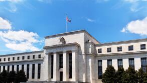 Exterior view of Federal Reserve Board of Governors Building facade entrance with eagle stone statue and American flags over a blue cloudscape sky.