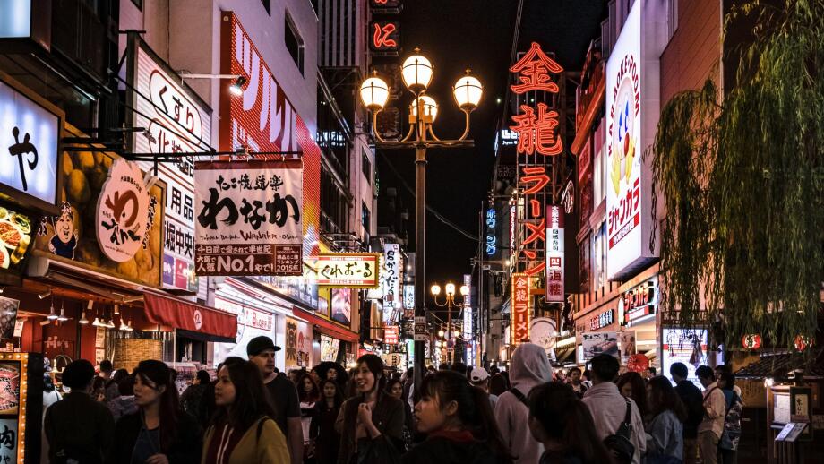 Crowd of tourists strolling down one of the streets in Osaka's Dotonbori area. Cityscape by night with lots of billboards and lights.