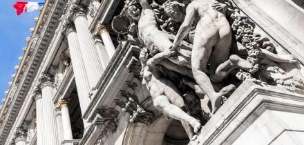 Close up of a group of marble statues on the facade of a french building, with a french flag waving in the background