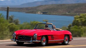 A 1958 Mercedes Benz 300SL Roadster driving along a scenic highway near a lake.