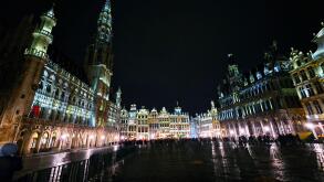 Night view of the illuminated Grand Place in Brussels, Belgium, with historic buildings and crowds gathered in the square.