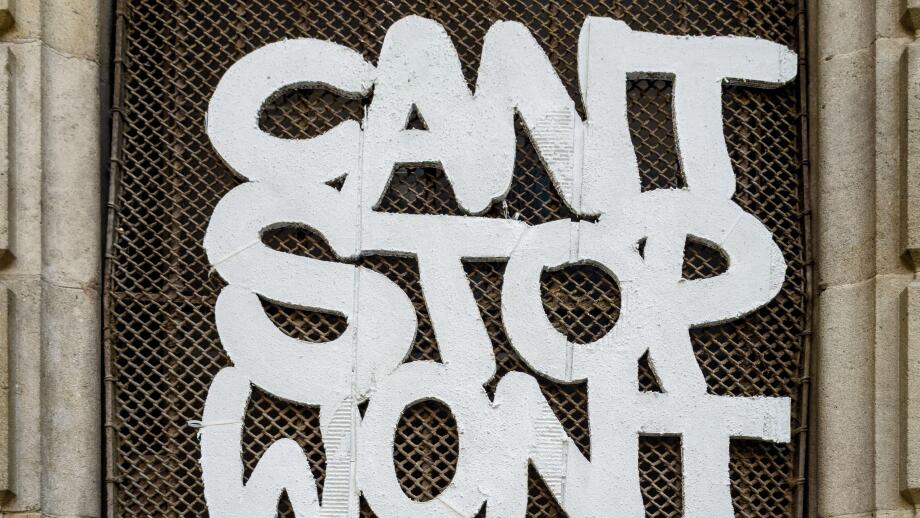 Graffiti with the message 'Can't Stop Won't Stop' on a wire mesh window grille in a historic stone doorway in Barcelona, Spain.