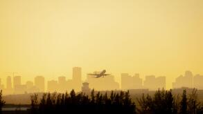 A plane is silhouetted against a colorful Yellow and Orange sunrise as it takes off from the Miami airport with the city in the background.