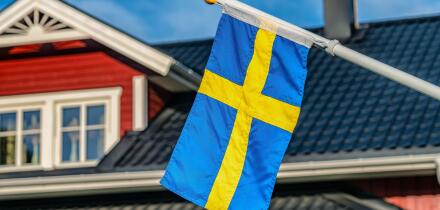 Close up photo of Swedish flag, blurry black roof and windows at top of Swedish wooden red house at background. Blue sky at sunny day