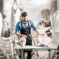 Portrait of a handsome sculptor in blue t-shirt and apron working with stone sculptures on the table at the old atmospheric studio