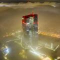 Aerial view of the European Central Bank skyscraper piercing through a sea of fog, its red lights ablaze against the muted city lights, Frankfurt am M