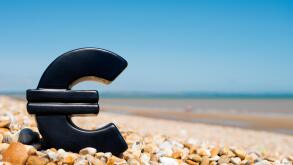 The Euro symbol sitting on a pebble beach against a clear blue sky.