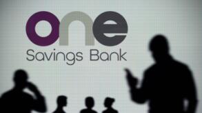 The OneSavings Bank logo is seen on an LED screen in the background while a silhouetted person uses a smartphone (Editorial use only)