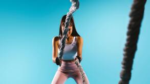 A strong, pumped-up girl in sport leggings throws a battle rope towards the camera. Blue background and empty side ad space.