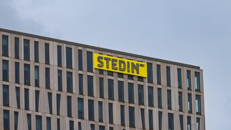 The top corner of the modern Stedin office building, showing its distinctive facade and the company's bright yellow logo against a grey sky. Rotterdam
