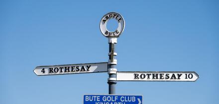 An island road sign on the Isle of Bute, Scotland against a blue sky. It shows the same destination, Rothesay, in two directions.