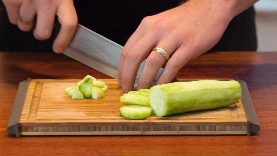 A male hand slicing a cucumber on a wooden cutting board.