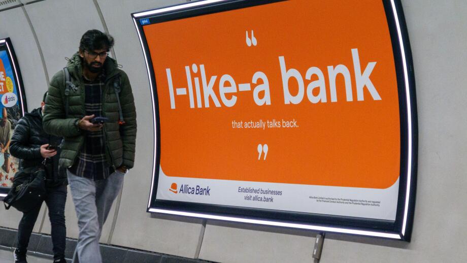 Commuters walk past an Advert for Allica Bank on wall of London railway station  London UK