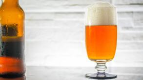 Beer glass full, with froth, and reflection, rugged stone wall as backdrop, and a beet bottle on the side, backlit