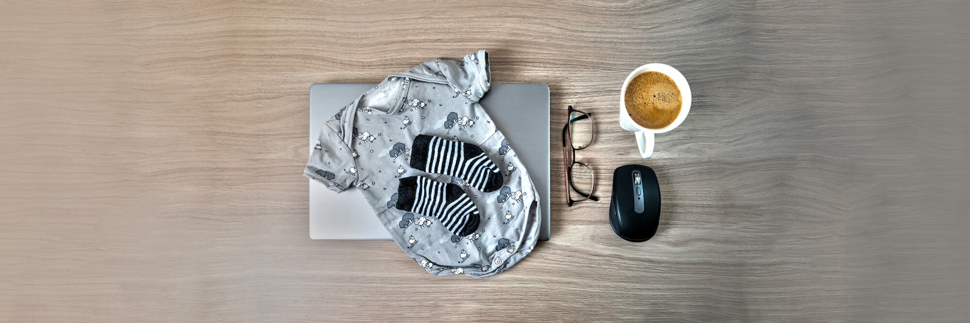 Baby socks, baby onesie, coffee, glasses laptop on wooden table 