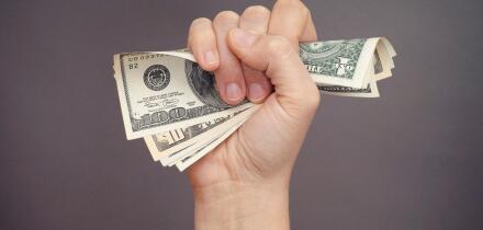 Woman hand squeezes dollar bills against grey background.
