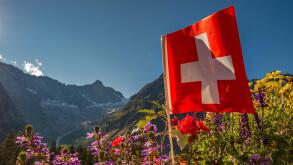 A bright red and white swiss flag flying in the wind with flowers and glacier covered peaks in the background in La Fouly, Switzerland
