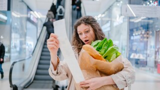A woman with a paper bag with groceries stands in a shopping center and looks in surprise at a check with high prices.