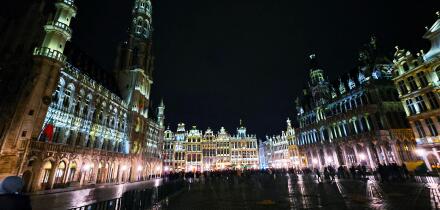 Night view of the illuminated Grand Place in Brussels, Belgium, with historic buildings and crowds gathered in the square.