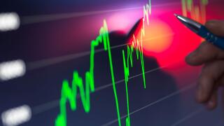 A hand holding a pen points to a bright stock market chart on a digital screen with fluctuating green and red graph lines indicating financial data.