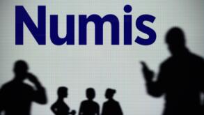 The Numis Securities logo is seen on an LED screen in the background while a silhouetted person uses a smartphone (Editorial use only)