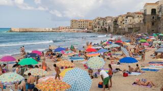 Cefalu, Sicily, Italy, crowds of people cover the sandy beach enjoying their holiday on a bright sunny August day with the old town buildings as a bac