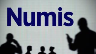 The Numis Securities logo is seen on an LED screen in the background while a silhouetted person uses a smartphone (Editorial use only)