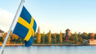 Swedish flag waving over calm waters in the Stockholm archipelago, with Djurgarden island and autumn foliage in the background on a peaceful early Oct