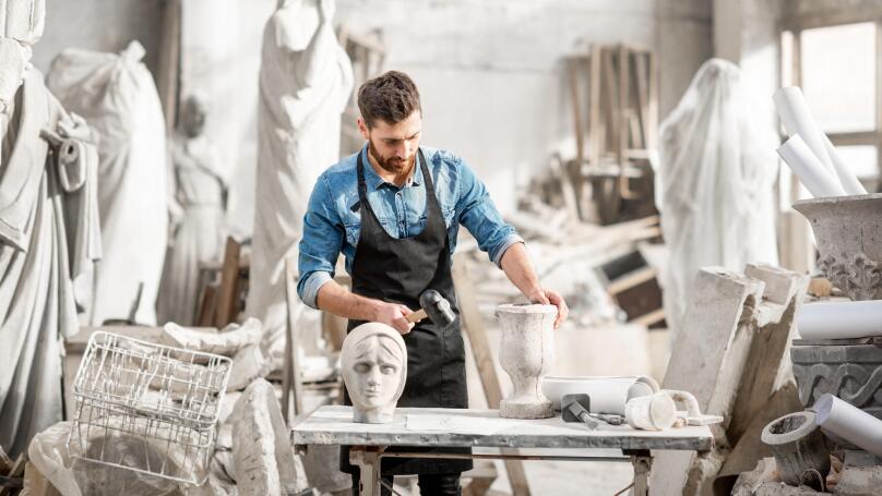Portrait of a handsome sculptor in blue t-shirt and apron working with stone sculptures on the table at the old atmospheric studio