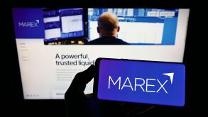 Person holding mobile phone with logo of British financial services company Marex Group on screen in front of web page. Focus on phone display.