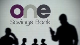 The OneSavings Bank logo is seen on an LED screen in the background while a silhouetted person uses a smartphone (Editorial use only)