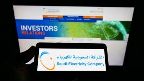 Person holding cellphone with logo of electric utility business Saudi Electricity Company (SEC) on screen with webpage. Focus on phone display.