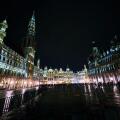 Night view of the illuminated Grand Place in Brussels, Belgium, with historic buildings and crowds gathered in the square.