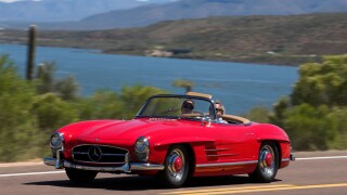 A 1958 Mercedes Benz 300SL Roadster driving along a scenic highway near a lake.