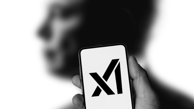 The logo of Elon Musk's xAI startup on a smartphone, being held up in front of a blurred image of the billionaire